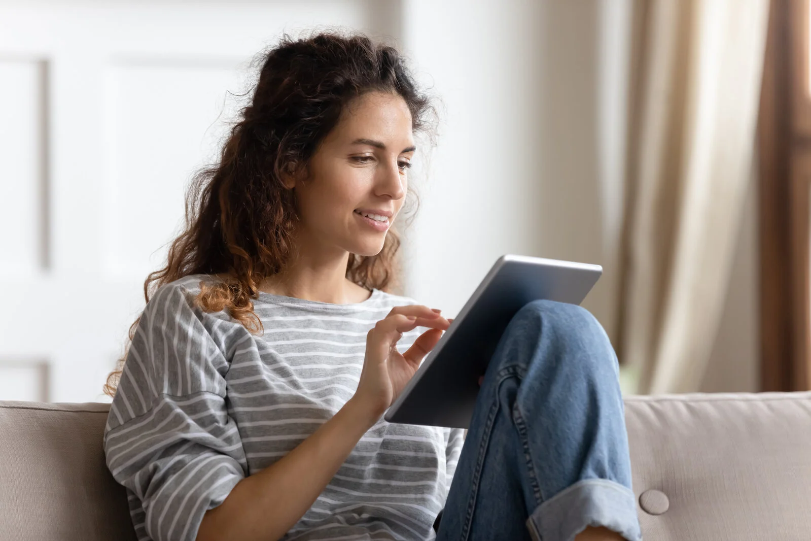 Smiling young woman using computer tablet, sitting on couch, typing on touchscreen, chatting in social network with friends, playing mobile device game or surfing internet, shopping online 2026/05/iStock-1257955550.jpg 